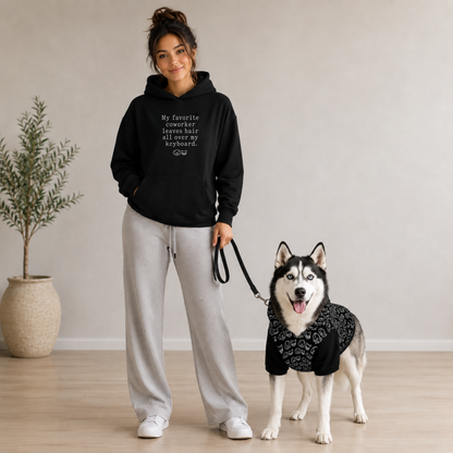 A woman stands indoors with a husky dog wearing a hoodie with white patterns. The woman wears a black hoodie with white text that reads, 'My favorite coworker leaves hair all over my keyboard.