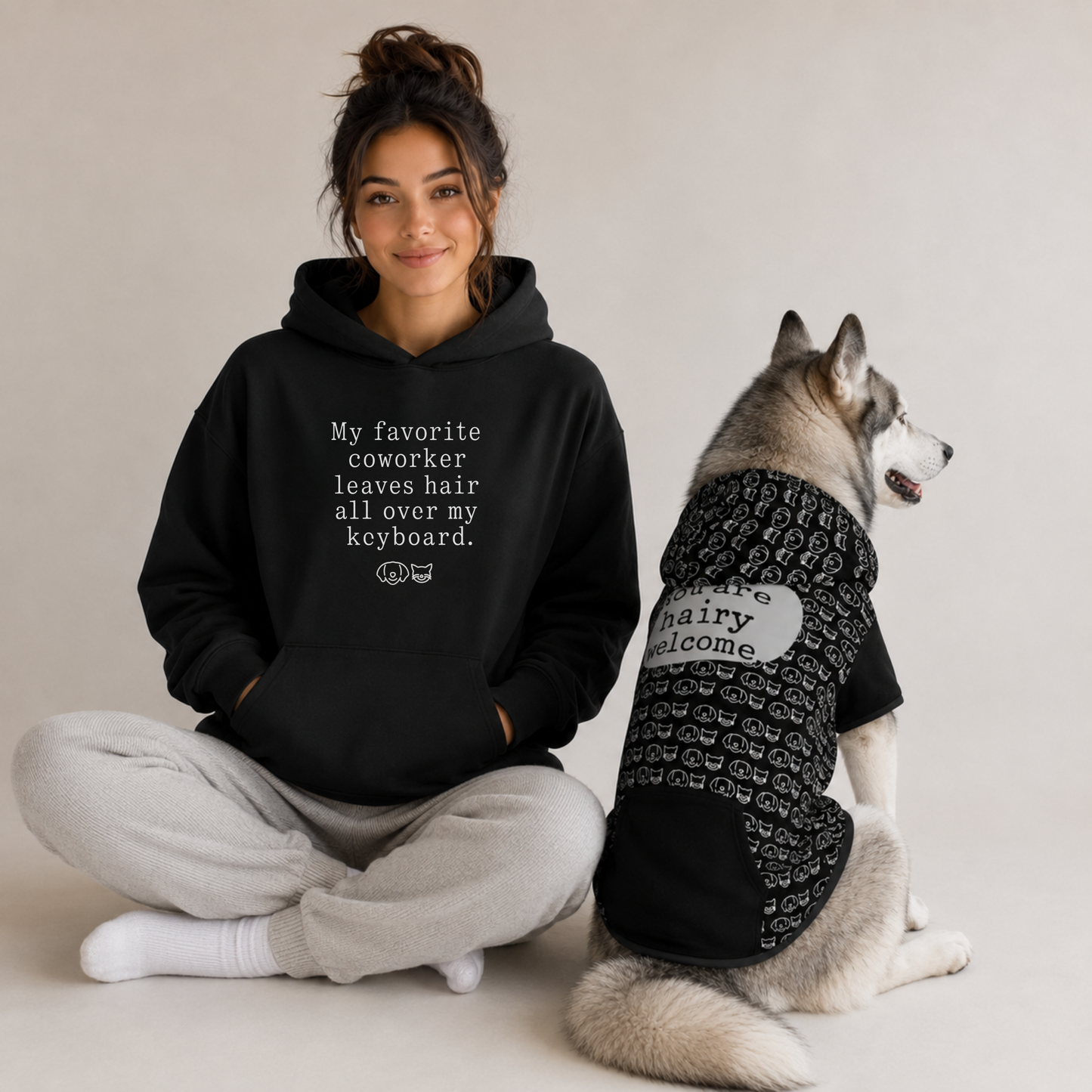 A woman sits cross-legged wearing a black hoodie with white text that reads, 'My favorite coworker leaves hair all over my keyboard.' beside a husky dog wearing a black hoodie with a white 'Hairy Welcome' patch.