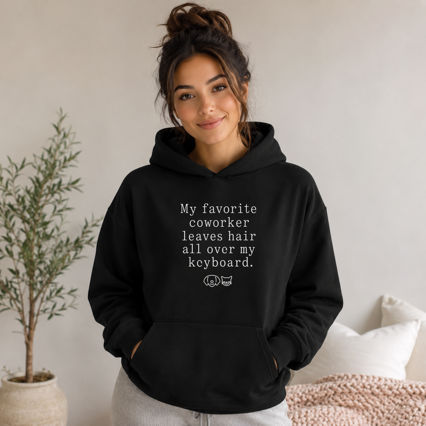 A woman wearing a black pullover hoodie with white text that reads 'My favorite coworker leaves hair all over my keyboard.' and a small cat graphic below, posed in a bright indoor room with a plant and bed visible.