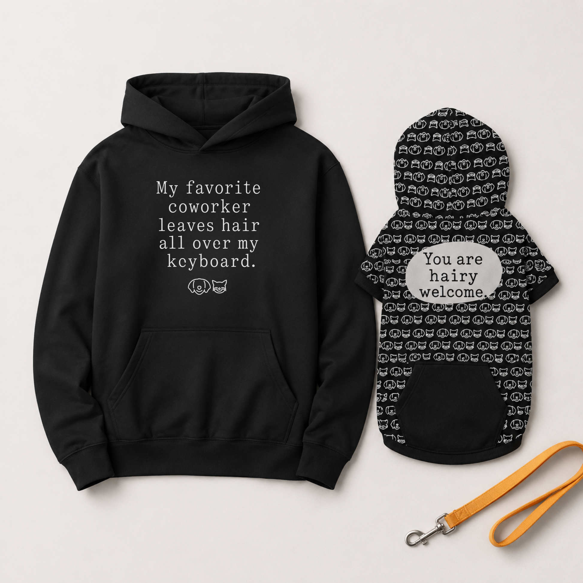 A black hoodie and matching black pet dog coat with white text and cat-face pattern, displayed on a light gray background with an orange leash.