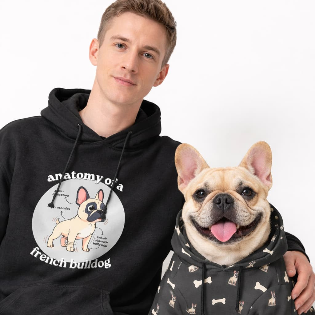 Man wearing a black hoodie with a dog graphic and text, sitting next to a dog in a matching black hoodie with bone patterns.