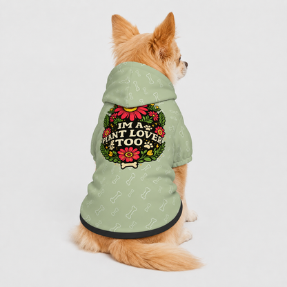 A small tan dog sits facing away from the camera wearing a sage green dog hoodie with a floral wreath graphic and the text 'I'M A PLANT LOVER TOO'.