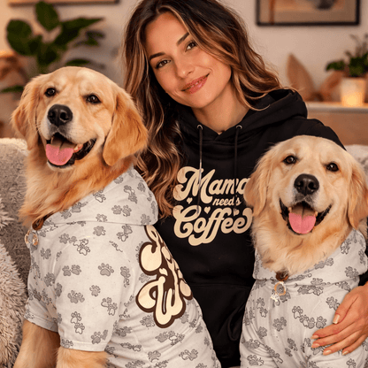 Woman sitting on a couch with two golden retrievers wearing matching pajamas, with a cup of coffee on a small table.