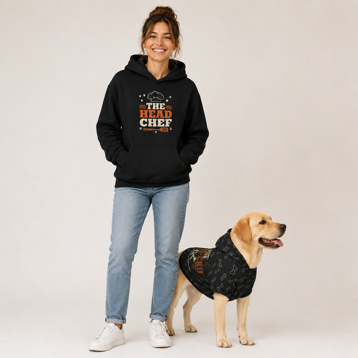 A woman wearing a black hoodie with a 'The Head Chef' graphic stands next to a yellow Labrador Retriever wearing a matching black dog hoodie, against a light gray studio background.