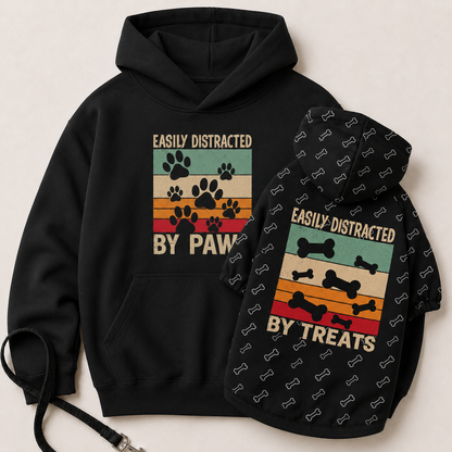 A black pet hoodie shown from both front and back on a light beige background. The front features a retro-style multicolor graphic with paw prints and the text 'EASILY DISTRACTED BY PAW'.