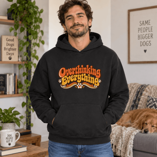 A man wearing a black hoodie with a graphic that reads 'Overthinking Everything' in orange and yellow, standing in a cozy living room with a dog on a couch in the background.