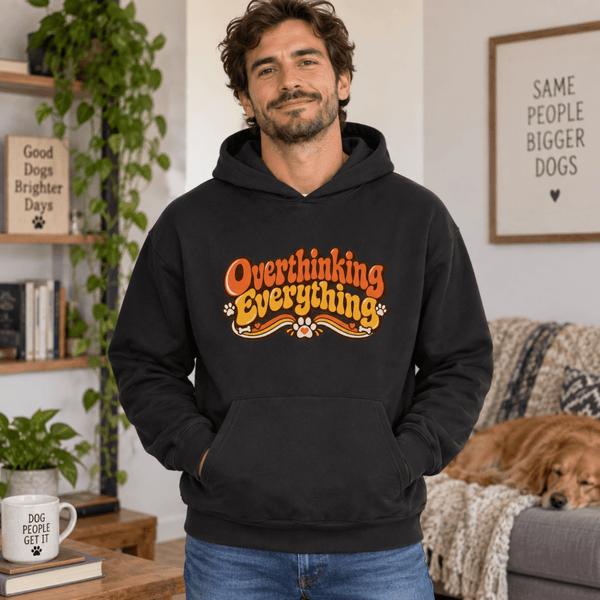 A man wearing a black hoodie with a graphic that reads 'Overthinking Everything' in orange and yellow, standing in a cozy living room with a dog on a couch in the background.