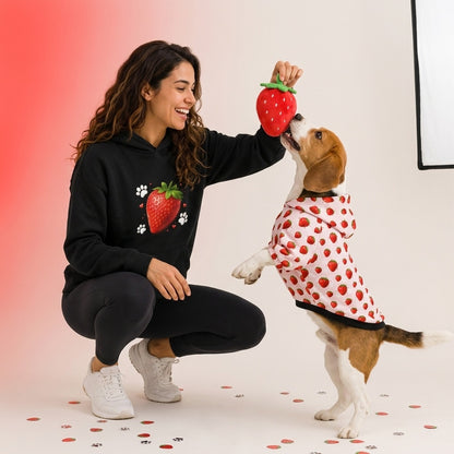 Woman in a black hoodie with strawberry design playing with a dog wearing a red and white polka dot outfit on a light background.
