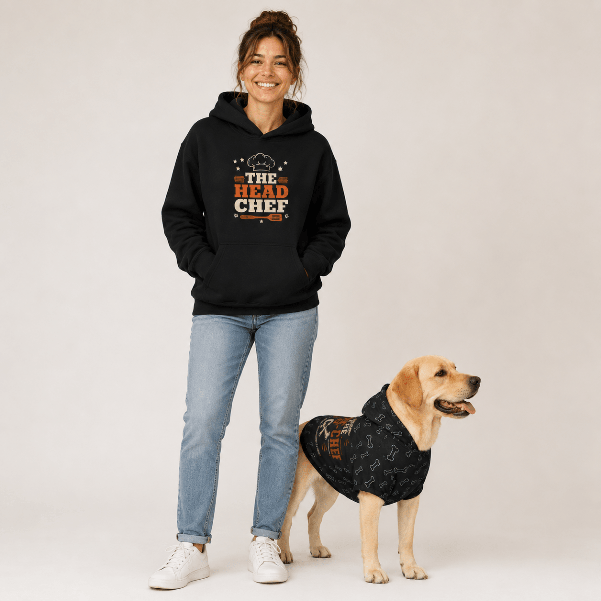 A woman wearing a black hoodie with a 'The Head Chef' graphic stands next to a yellow Labrador Retriever wearing a matching black dog hoodie, against a light gray studio background.
